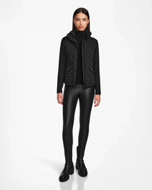 A woman faces forward in Rudsaks KARMA - L124503 Black quilted vest for sustainable warmth over a turtleneck, styled with faux leather leggings and ankle boots—an ideal transitional weather look—against a plain white background.