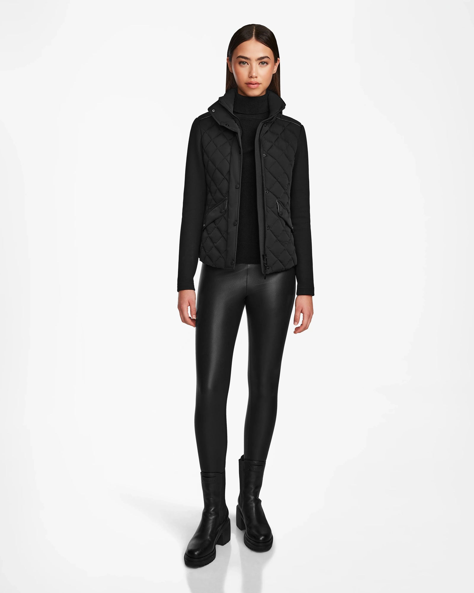 A woman faces forward in Rudsaks KARMA - L124503 Black quilted vest for sustainable warmth over a turtleneck, styled with faux leather leggings and ankle boots—an ideal transitional weather look—against a plain white background.