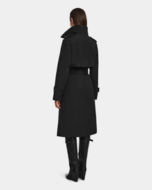A woman stands facing away, wearing the Rudsak KINDA - 8124690 Black womens trench coat with leather detailing, a high collar, embossed lettering at the back, waist belt, and knee-high black boots.