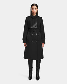 A woman stands facing forward against a plain white background, wearing the Rudsak KINDA - 8124690 Black womens trench coat with leather details, belted waist, long sleeves, and black knee-high boots.