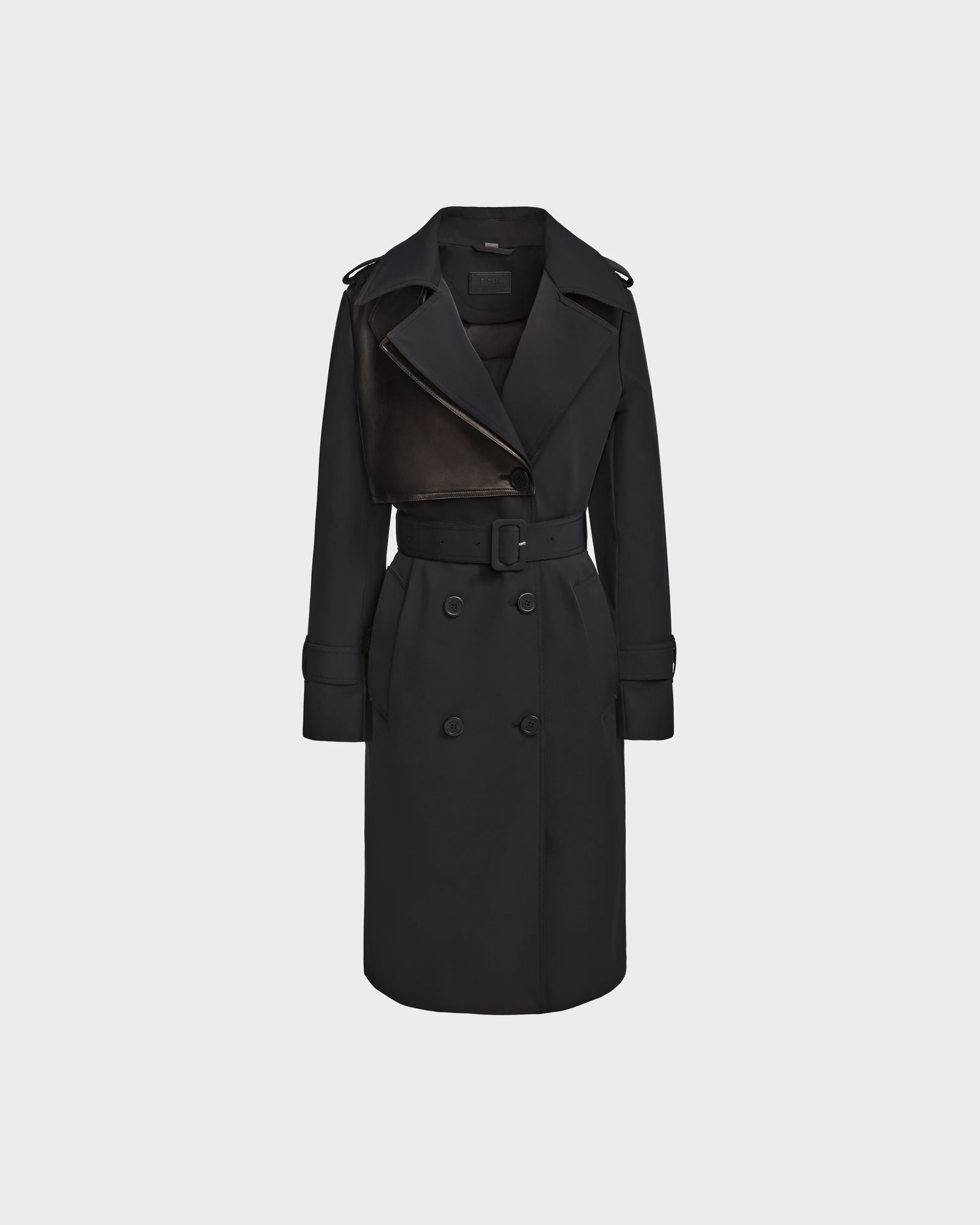 The KINDA - 8124690 Black by Rudsak is a womens long black trench coat with leather accents, double-breasted front, belted waist, wide lapels, and buttoned cuffs, shown on a plain light gray background.