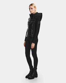 A person stands slightly sideways against a plain white background, wearing the RUDSAK MABS - 8122020 BLACK womens puffer jacket with RDS-certified down, paired with black leggings and black lace-up boots.