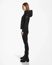 A woman with straight brown hair stands in profile against a white background, wearing the Rudsak MALYA M - L123547 Black down quilted jacket, paired with black leggings and boots for an activewear-inspired look.