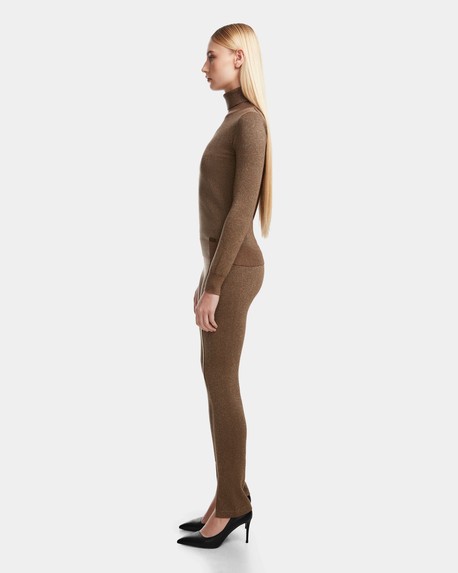 A woman with long straight blonde hair stands in profile, wearing Rudsak MANSUR - 8524624 Antique Bronze, paired with chic ankle slit leggings and black high heels, against a plain white background.