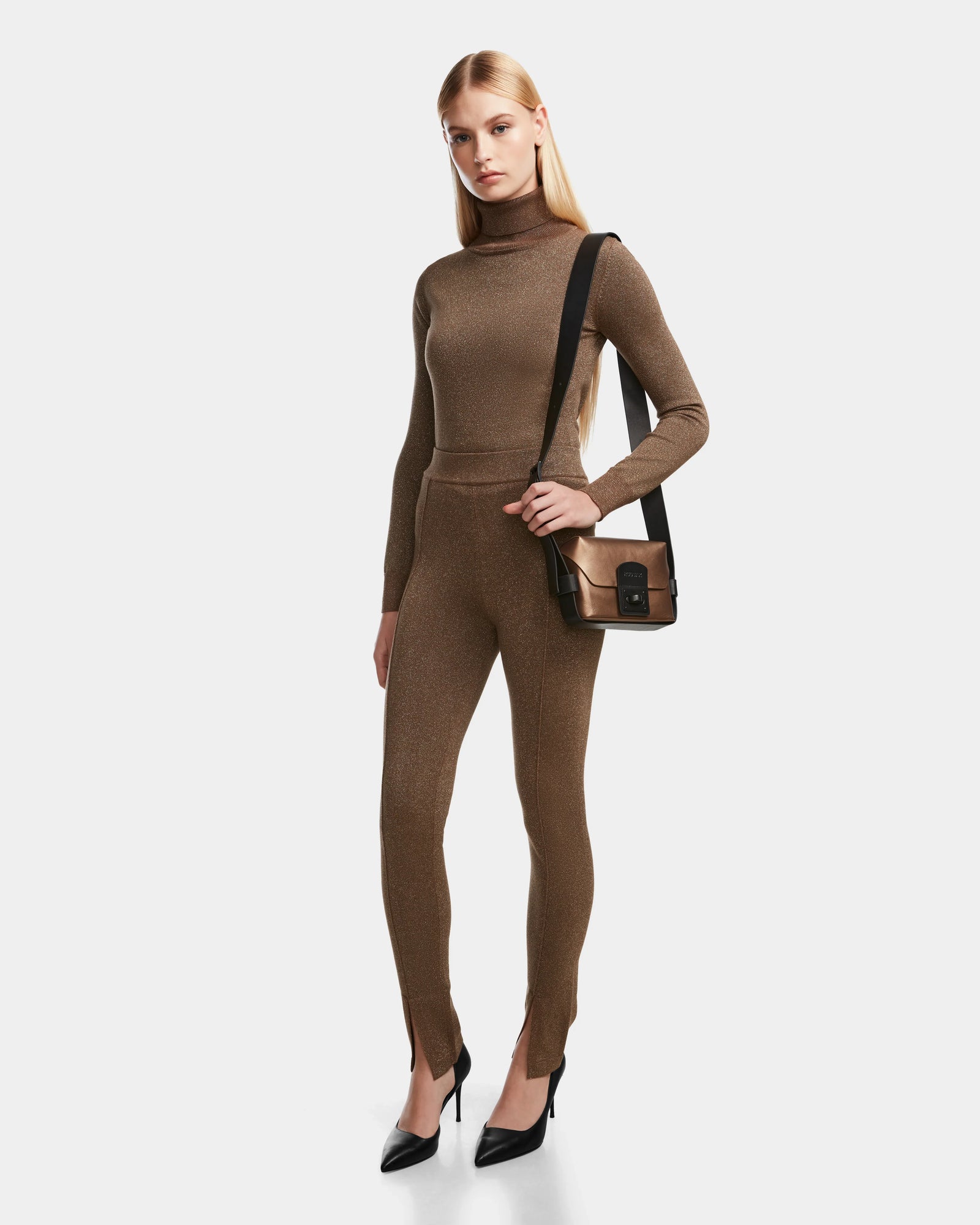 A woman with long blonde hair wears a fitted brown turtleneck, ankle slit leggings, black heels, and carries the Rudsak MANSUR - 8524624 Antique Bronze crossbody bag with a black strap, standing against a plain white background.
