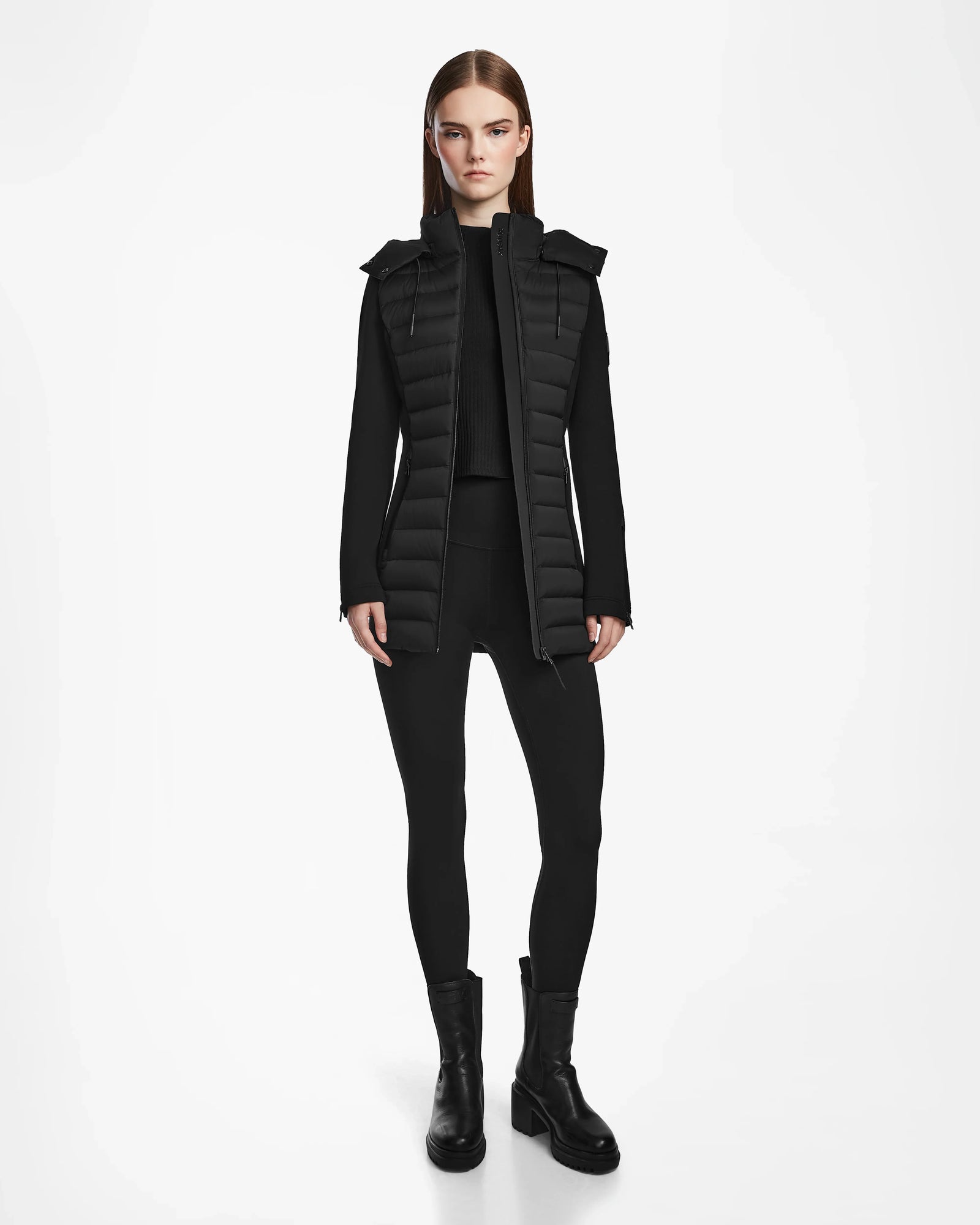 A woman stands facing forward in the MARRY M - L123548 Black puffer jacket by Rudsak, featuring a removable hood, worn over a long-sleeve top with black leggings and chunky boots against a plain white background.