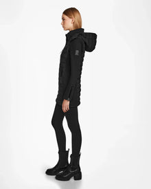 Woman in profile wearing the RUDSAK MERINA - 8122546 BLACK quilted down jacket with black leggings and ankle boots, standing against a plain white background.