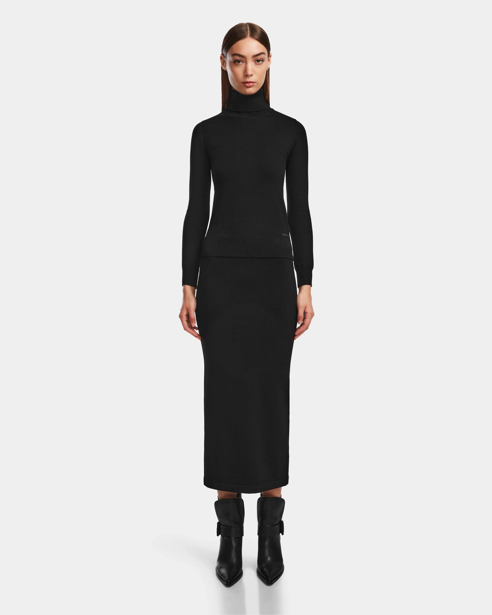 A woman stands facing forward against a plain white background, wearing an elegant black turtleneck sweater, the Rudsak MONICA - 8524516 Black womens knit skirt, and black ankle boots.