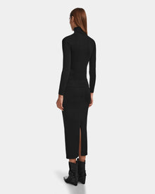 A woman with straight, long brown hair is seen from behind in a fitted black turtleneck and the MONICA - 8524516 Black knit skirt by Rudsak, which features a back slit. She pairs it with black ankle boots with bows against a plain white background.