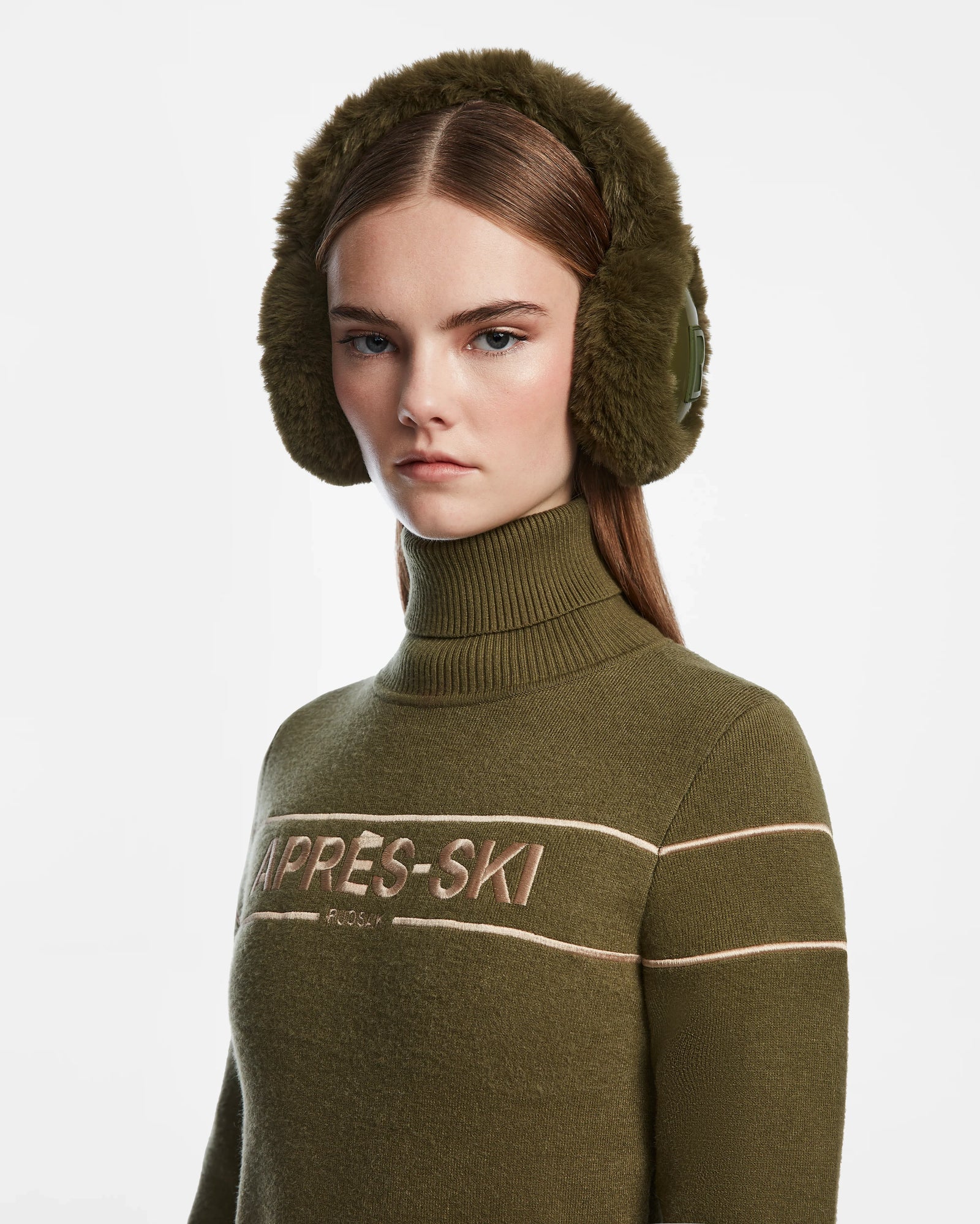 A woman with straight brown hair showcases Rudsak MUFF - 8422503 Loden Green faux fur earmuffs and an olive green “APRES-SKI” turtleneck against a white background.