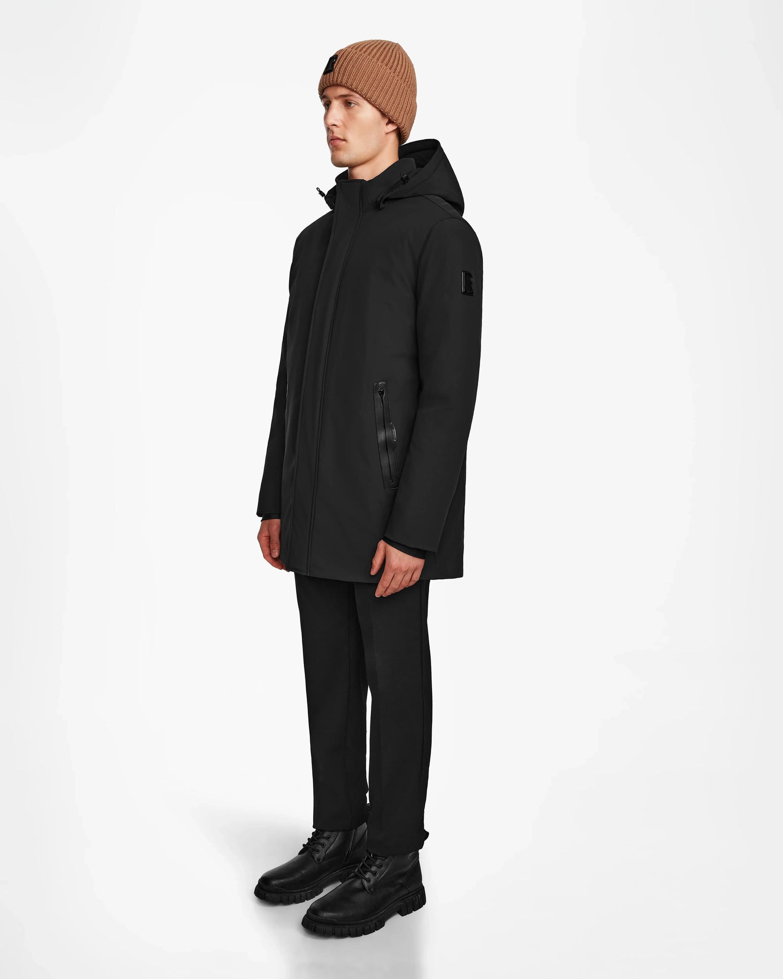 A man wearing the Rudsak NATAN - M124519 Black men’s parka with down fill, along with a brown beanie, black pants, and boots, stands against a plain white background, facing slightly to the side.