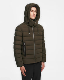 A person wearing the Rudsak NAVAN - 6124551 Army mens olive green hooded light down puffer with front zipper and zippered side pocket stands against a plain white background.