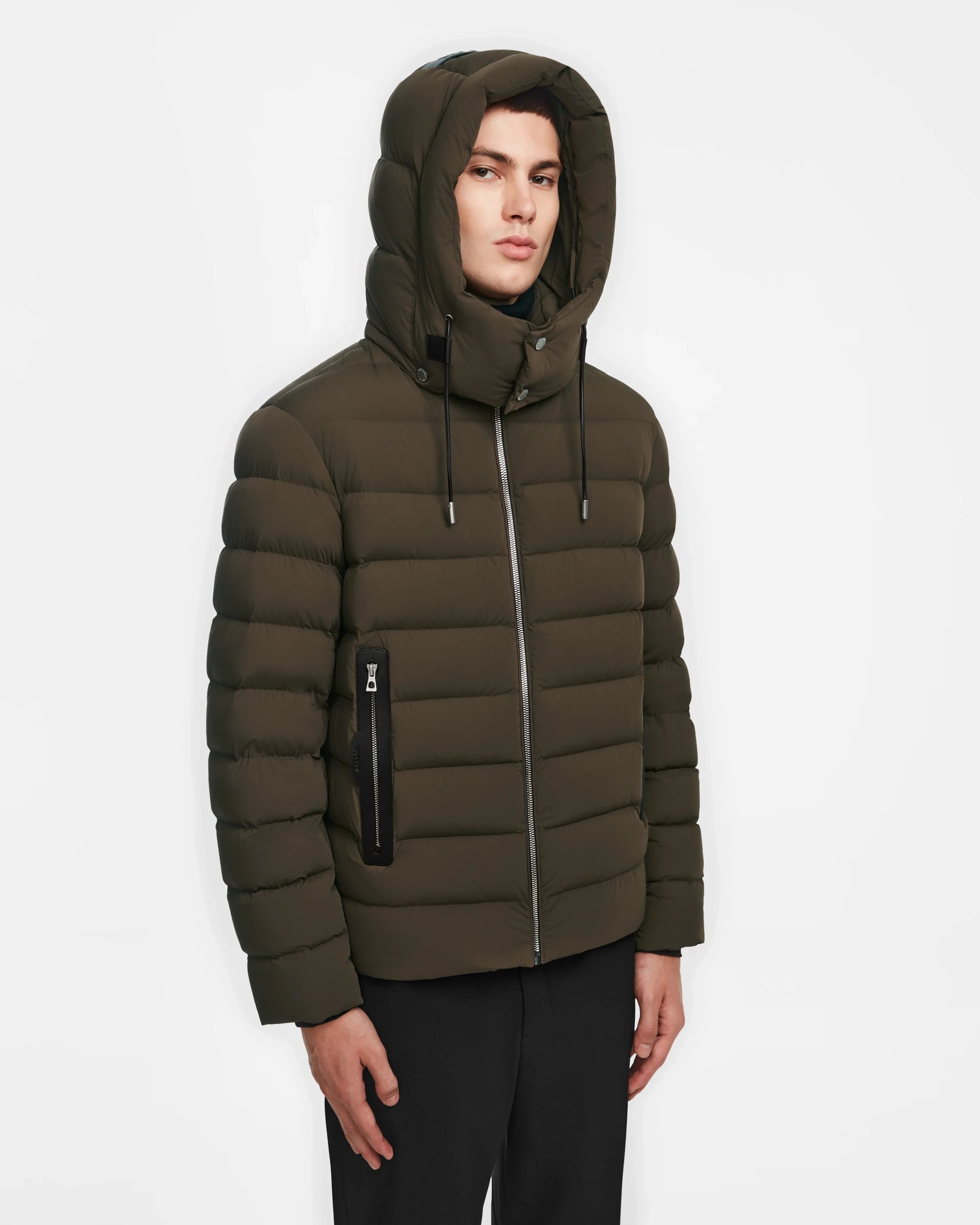 A person wearing the Rudsak NAVAN - 6124551 Army mens olive green hooded light down puffer with front zipper and zippered side pocket stands against a plain white background.