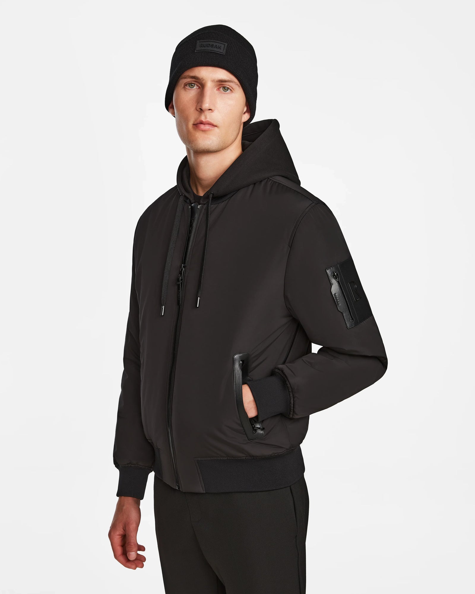 A man wearing the Rudsak NEVEN - M124520 Black men’s bomber jacket, made from recycled materials with zip closure and front pockets, black beanie, and black pants stands against a plain white background.
