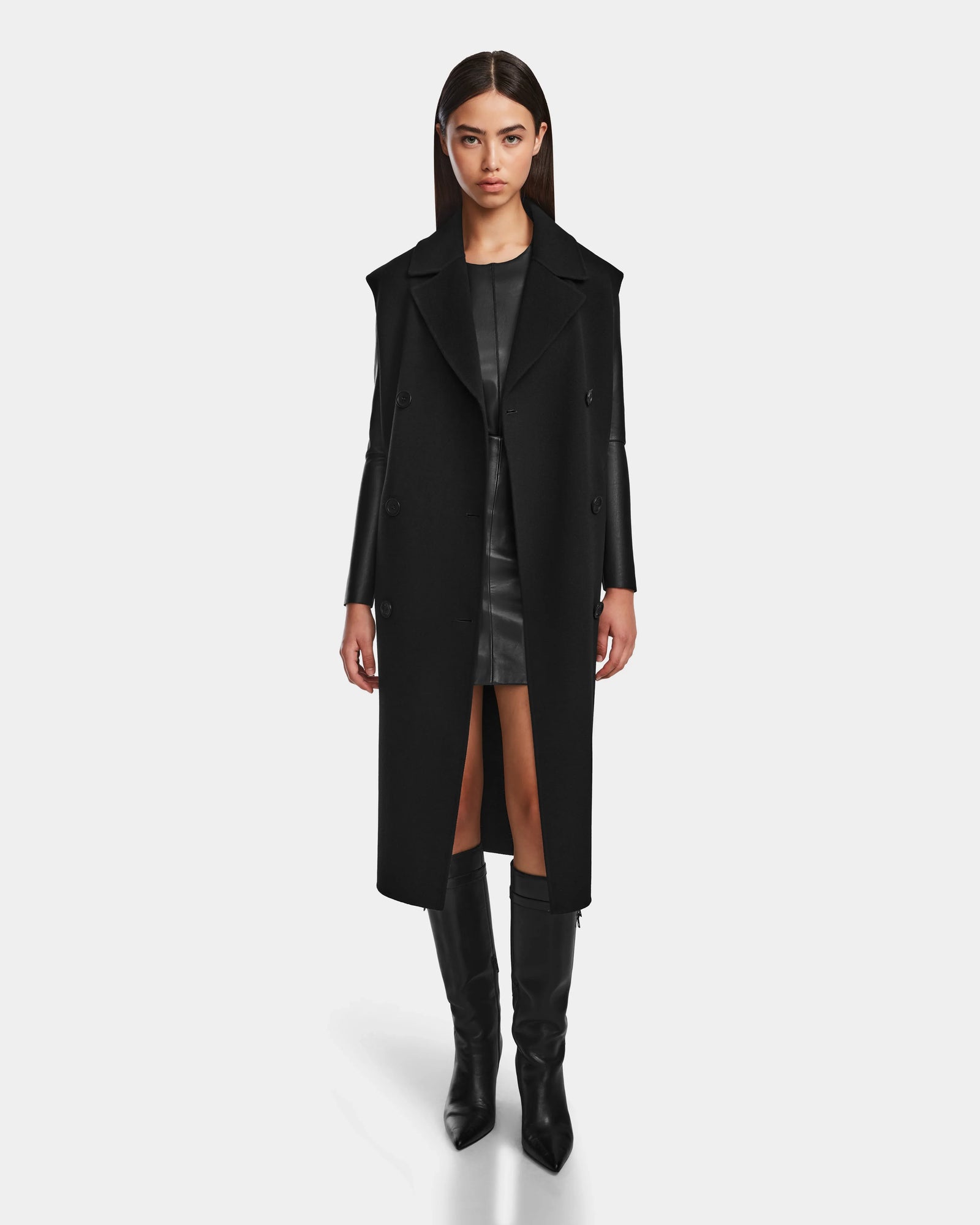 A woman with straight brown hair wears the Rudsak NOURA - 8124698 Black long coat over a wool vest, paired with a short black dress and knee-high boots, standing against a plain white background.