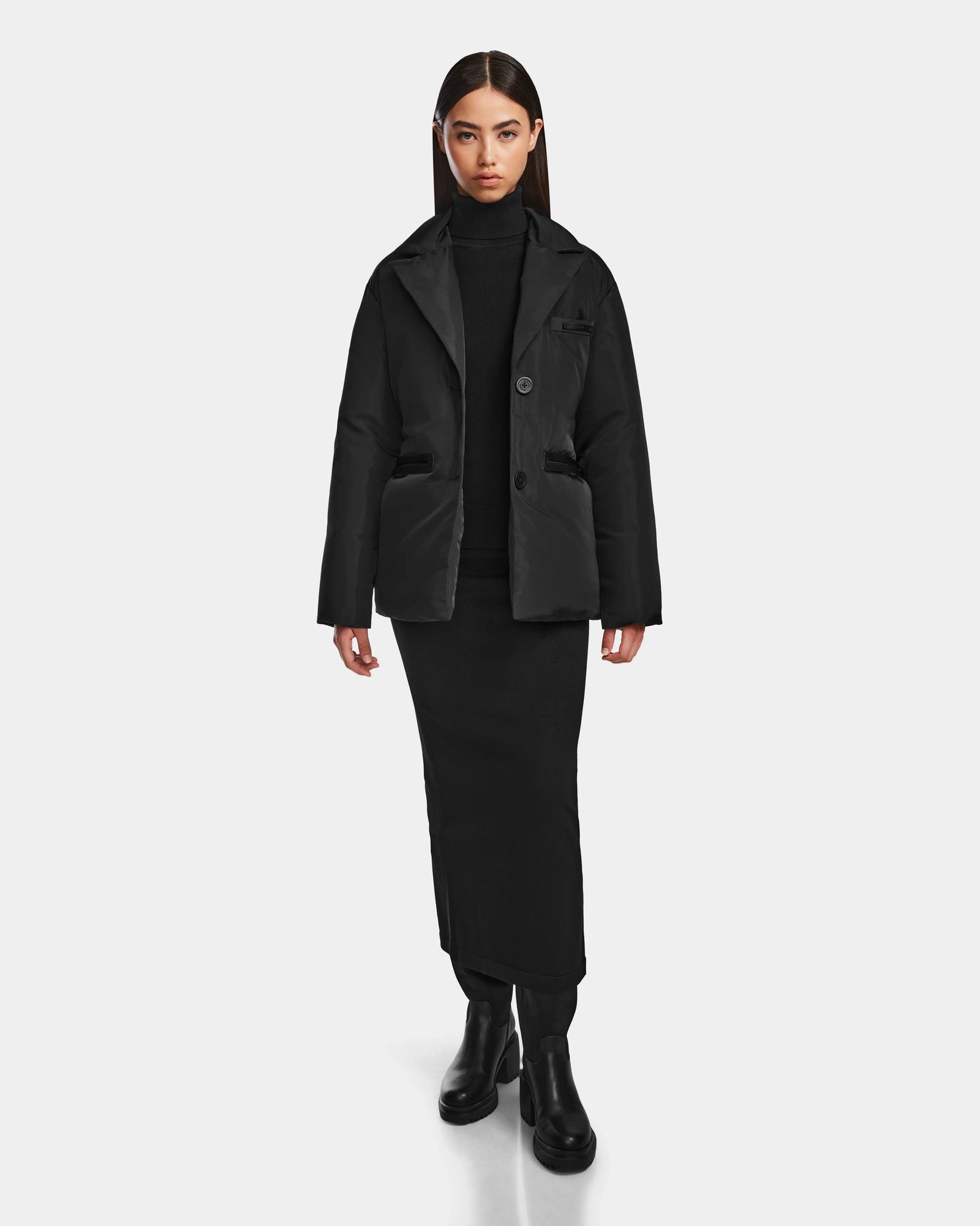 A woman faces forward in a Rudsak PAOLA - 8124547 Black weather-resistant puffer jacket, styled with a black turtleneck, long black skirt, and black boots against a plain white background.