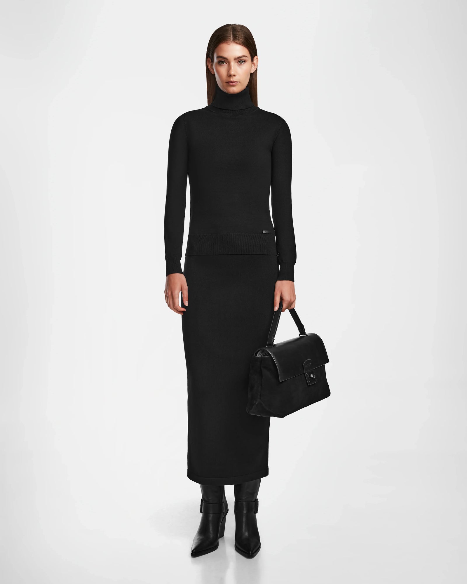 A woman stands against a white background wearing a fitted black turtleneck, long black skirt, black boots, and holding the Rudsak ESMA - 8324519 Black suede and leather handbag with a removable shoulder strap.