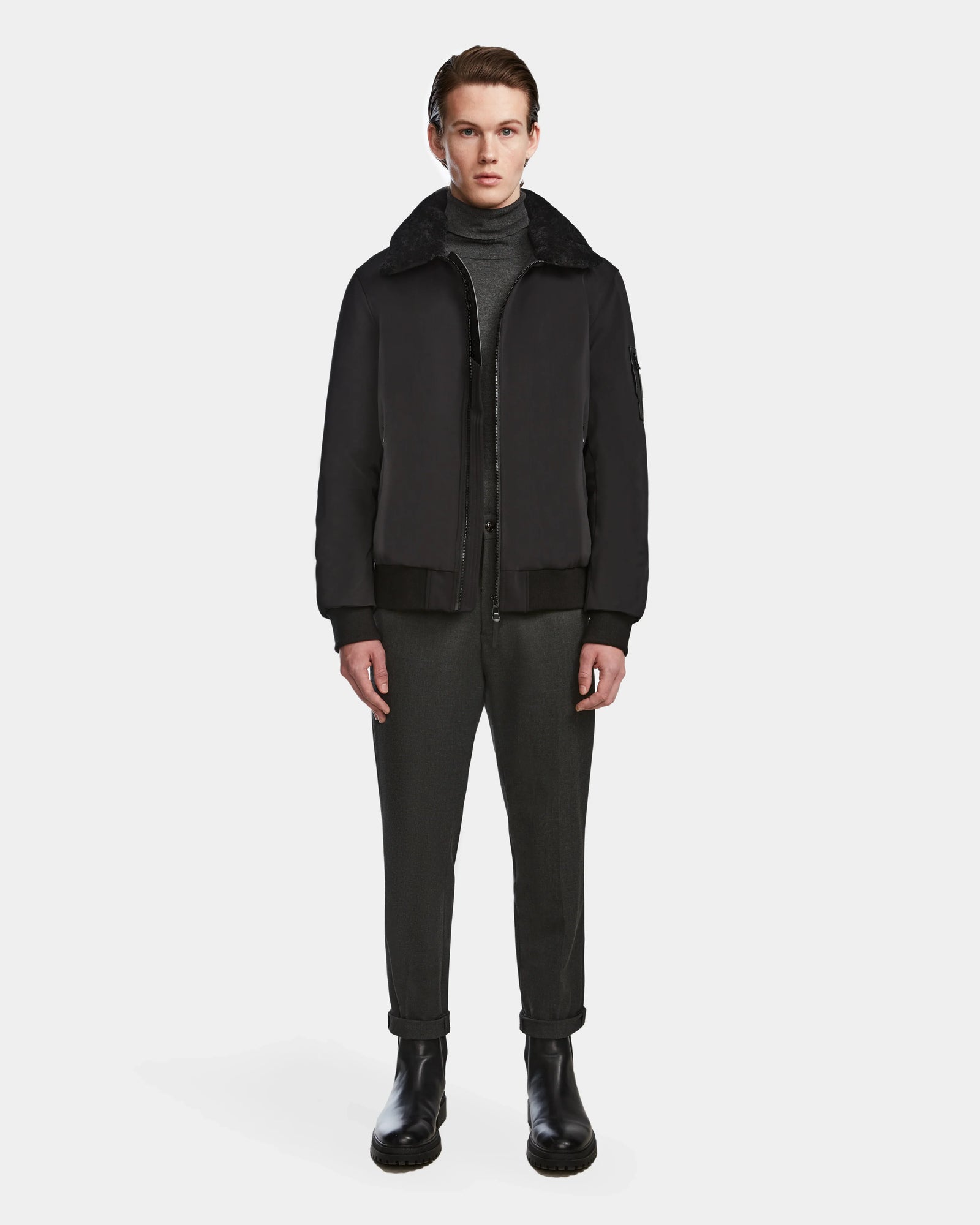 A person faces forward wearing the Rudsak REECE - 6124592 Black men’s bomber jacket with a shearling collar over a dark turtleneck, dark pants, and black boots against a plain white background.
