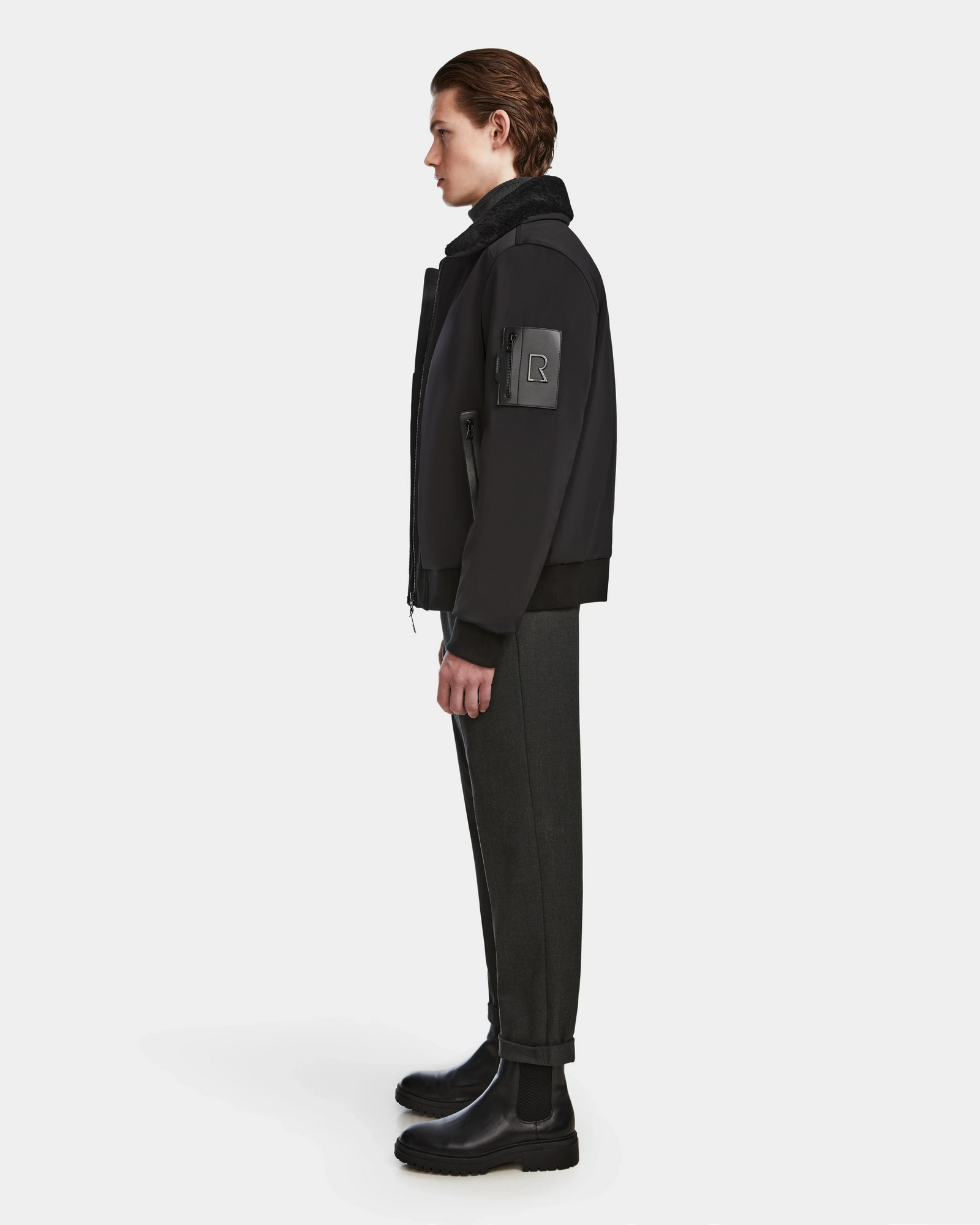 A young man with slicked-back hair stands in profile, wearing the Rudsak REECE - 6124592 Black water-resistant mens bomber jacket with a logo patch on the sleeve, paired with dark cuffed trousers and black boots against a white background.