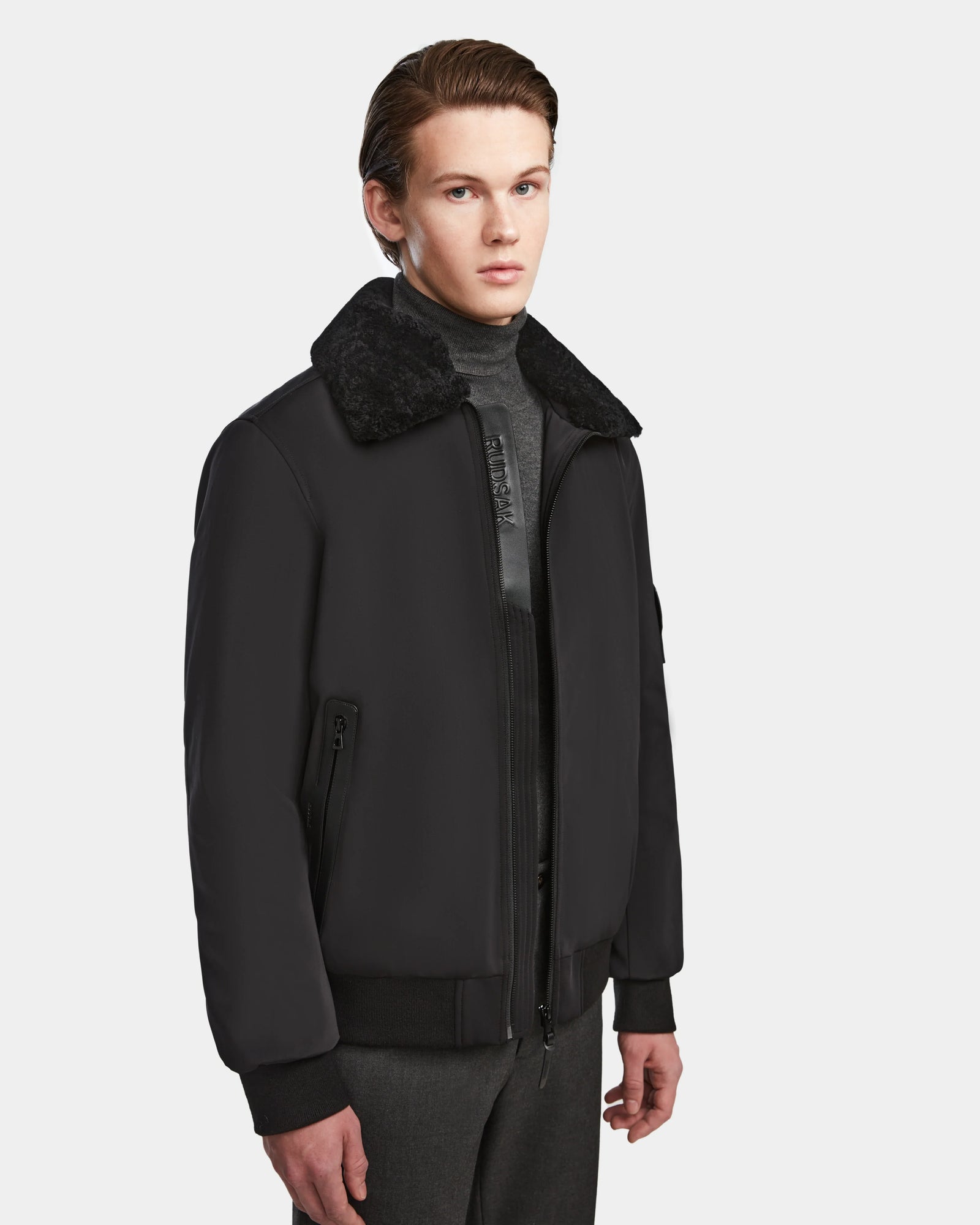A young man with short brown hair wears the Rudsak REECE - 6124592 Black men’s bomber jacket, featuring a dark shearling collar, over a gray turtleneck and gray pants, standing against a plain white background.