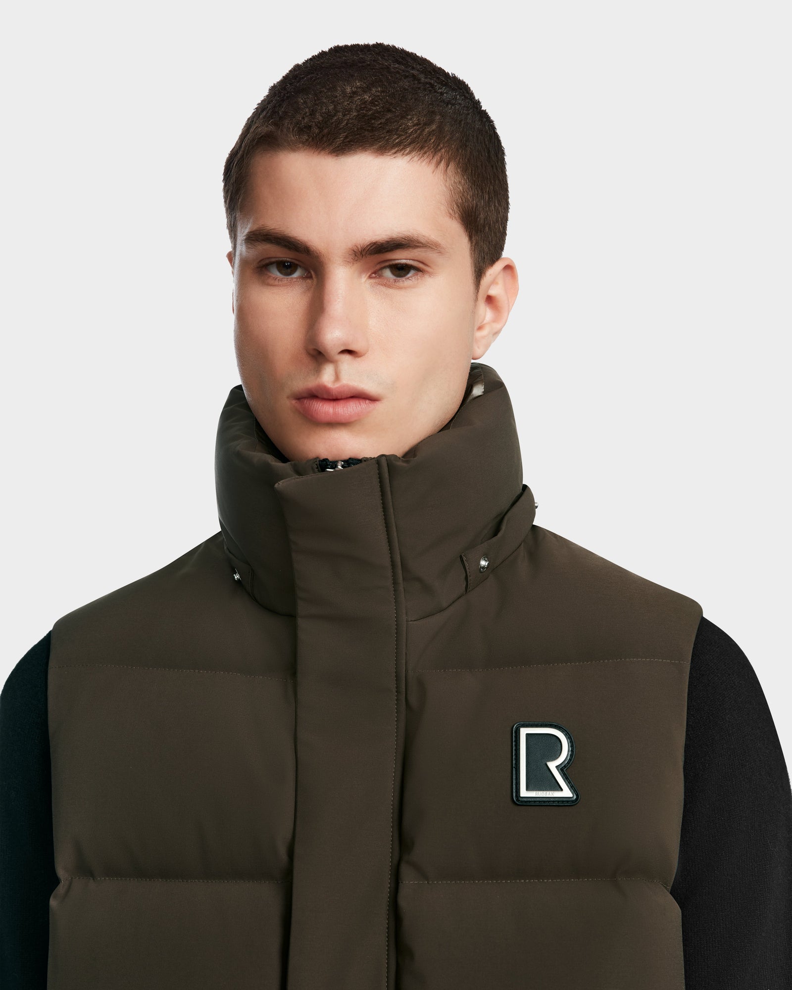 A young man with short brown hair wears the Rudsak REN M - 9123512 Army M high-collar dark green puffer vest, featuring a black R logo on the chest, layered over a black top against a plain light gray background.