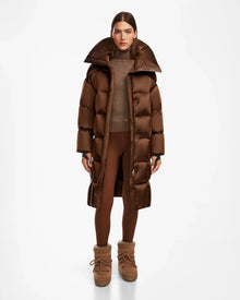 A woman stands in a studio wearing the Rudsak SASH R - 8124543 Antique Bronze womens long down puffer coat, paired with a brown sweater, brown leggings, and matching winter boots. She faces forward with a neutral expression against a white background.