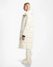 A woman in profile wears the SASH - 8124536 Iceland long, puffy womens down puffer coat by Rudsak with tall padded white boots against a plain white background.