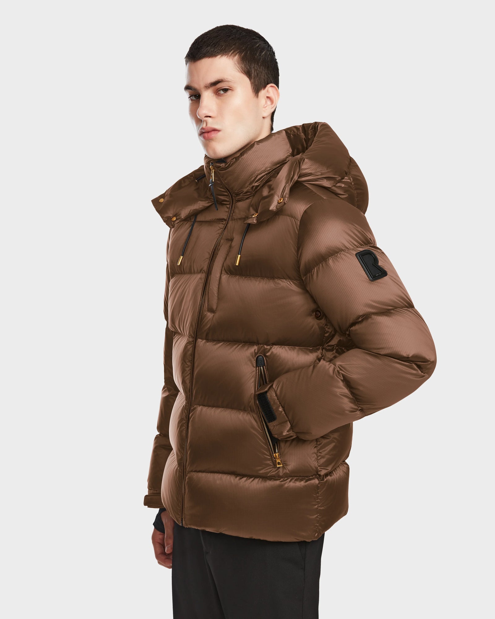 A young man wears the Rudsak VINCE S - 6124560 Antique Bronze mens quilted down puffer with a hood and RDS-certified down, standing against a plain white background with one hand in his pocket, looking slightly to the side.