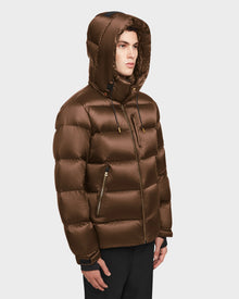 A young man wearing the Rudsak VINCE S - 6124560 Antique Bronze men’s quilted down puffer with a shiny finish, hood, and zippered pockets stands against a light background, facing slightly to the side.