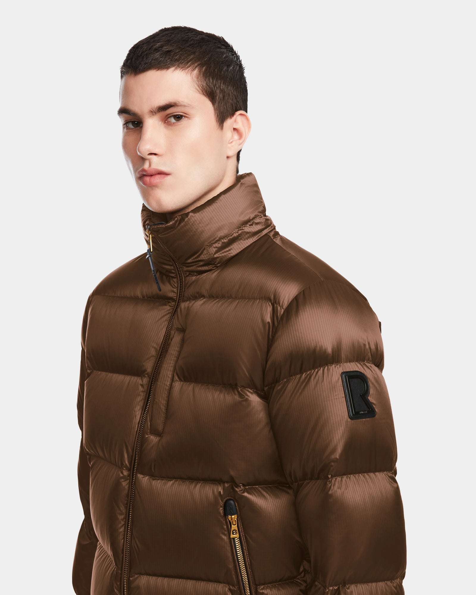 A young man with short dark hair wears the Rudsak VINCE S - 6124560 Antique Bronze men’s quilted down puffer jacket, featuring RDS-certified down and a black R patch on the sleeve, looking slightly to the side against a white background.