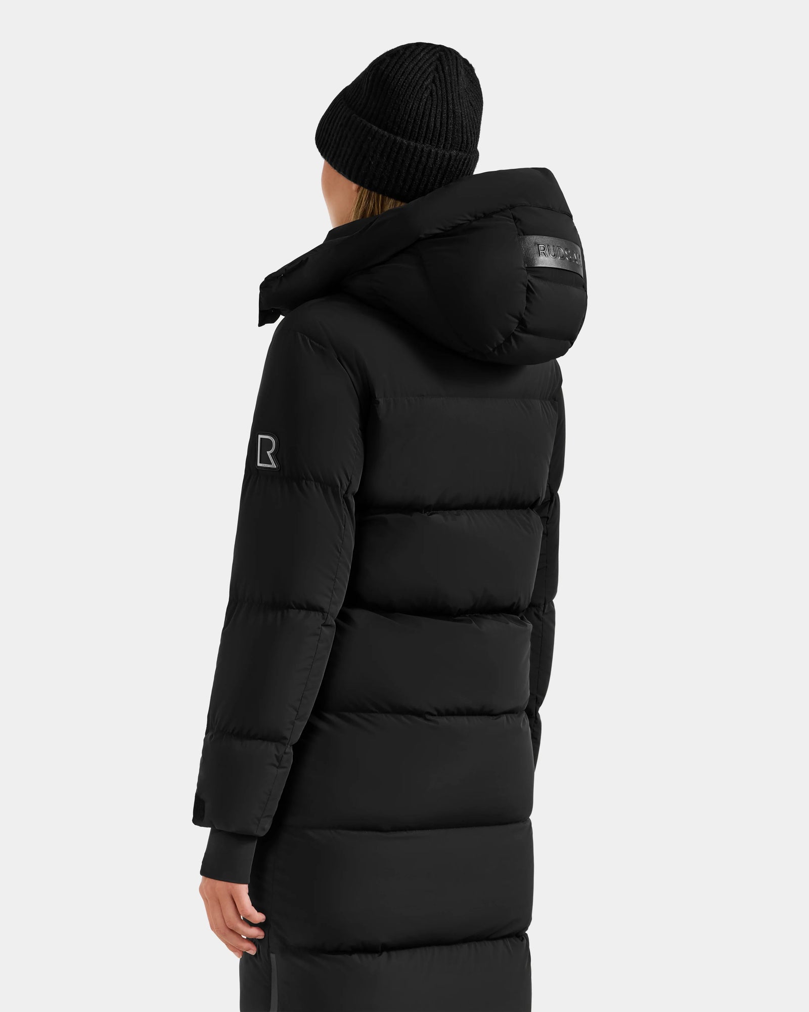 A person wears the Rudsak ALESSANDRA L - L125553 Black quilted down puffer coat with a hood and a black knit beanie, shown from the back against a plain light background—a true winter essential.