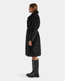 A woman in profile models the Rudsak ANNABELLE FSU - L125512 Black, a knee-length sherpa teddy trench coat with a belt, paired with black knee-high boots against a plain light background.