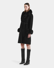 A woman with straight brown hair wears the Rudsak ARTIS - L125575 Black women’s wool coat featuring a faux fur collar and cuffs, belted at the waist, paired with black knee-high boots against a plain white background.