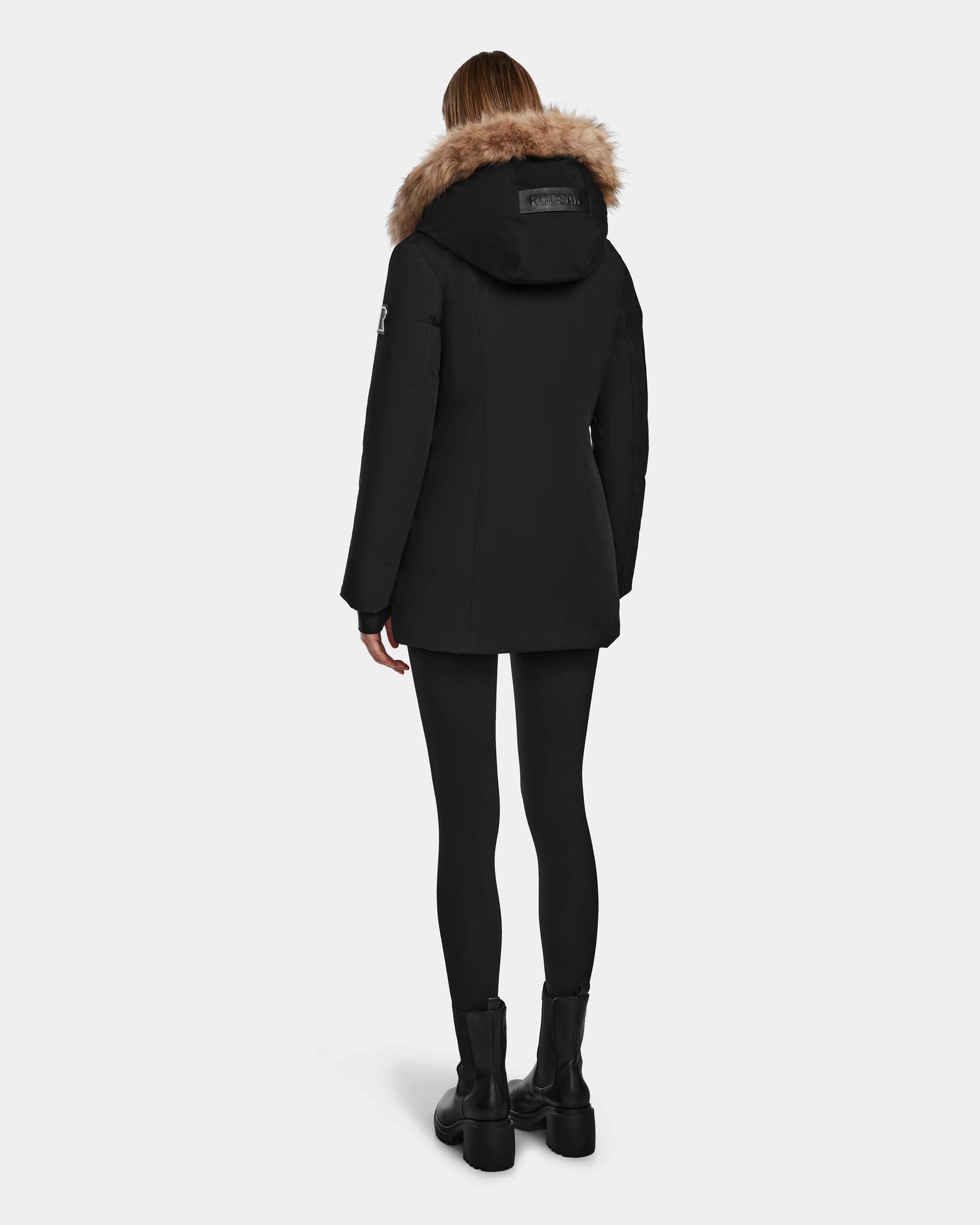 A person wearing the Rudsak AURORE - L123561 Black/Natural coat stands facing away, showcasing its fitted black design with fur trim against a plain light background.