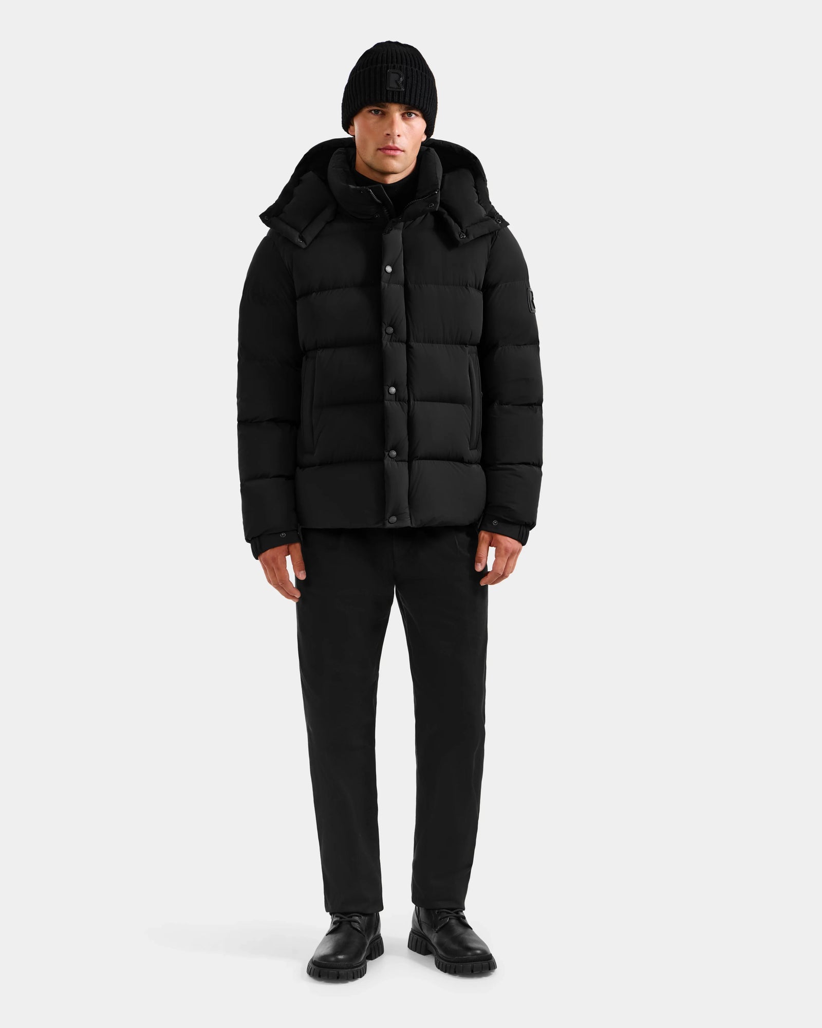 A person stands facing forward wearing the Rudsak BRAYDEN M-S - M125524 Black RDS-certified down puffer jacket, paired with a black beanie, pants, and shoes against a plain light background.