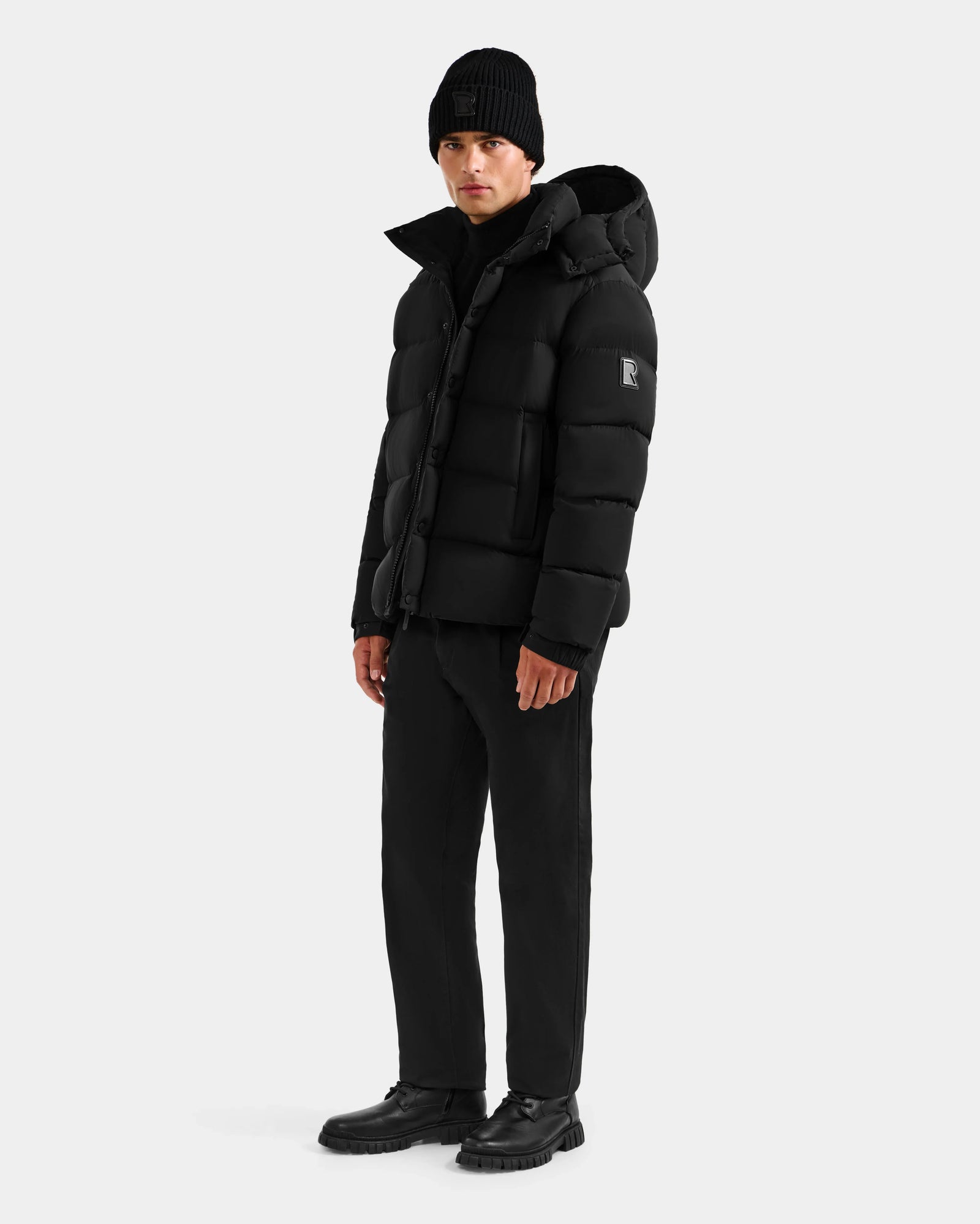 A man wearing the Rudsak BRAYDEN M-S - M125524 Black puffer jacket, black knit beanie, black pants, and black shoes stands against a plain light gray background.