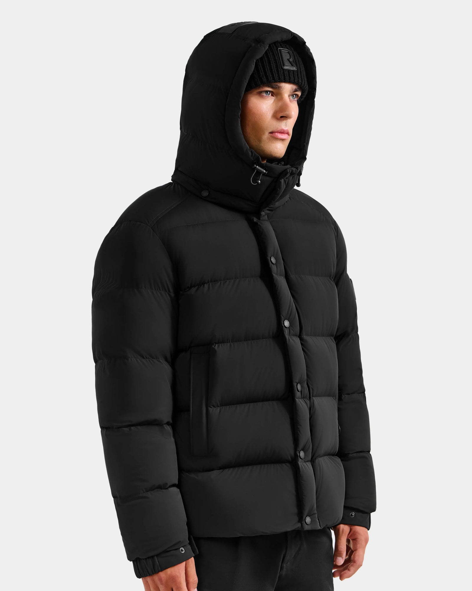 A man wearing the Rudsak BRAYDEN M-S - M125524 Black down puffer jacket and a black knit beanie stands against a plain light gray background.