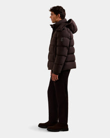A man stands in profile wearing the Rudsak BRAYDEN M-S - M125524 Espresso men’s down puffer jacket with a hood, paired with dark pants and shoes, against a plain light background.