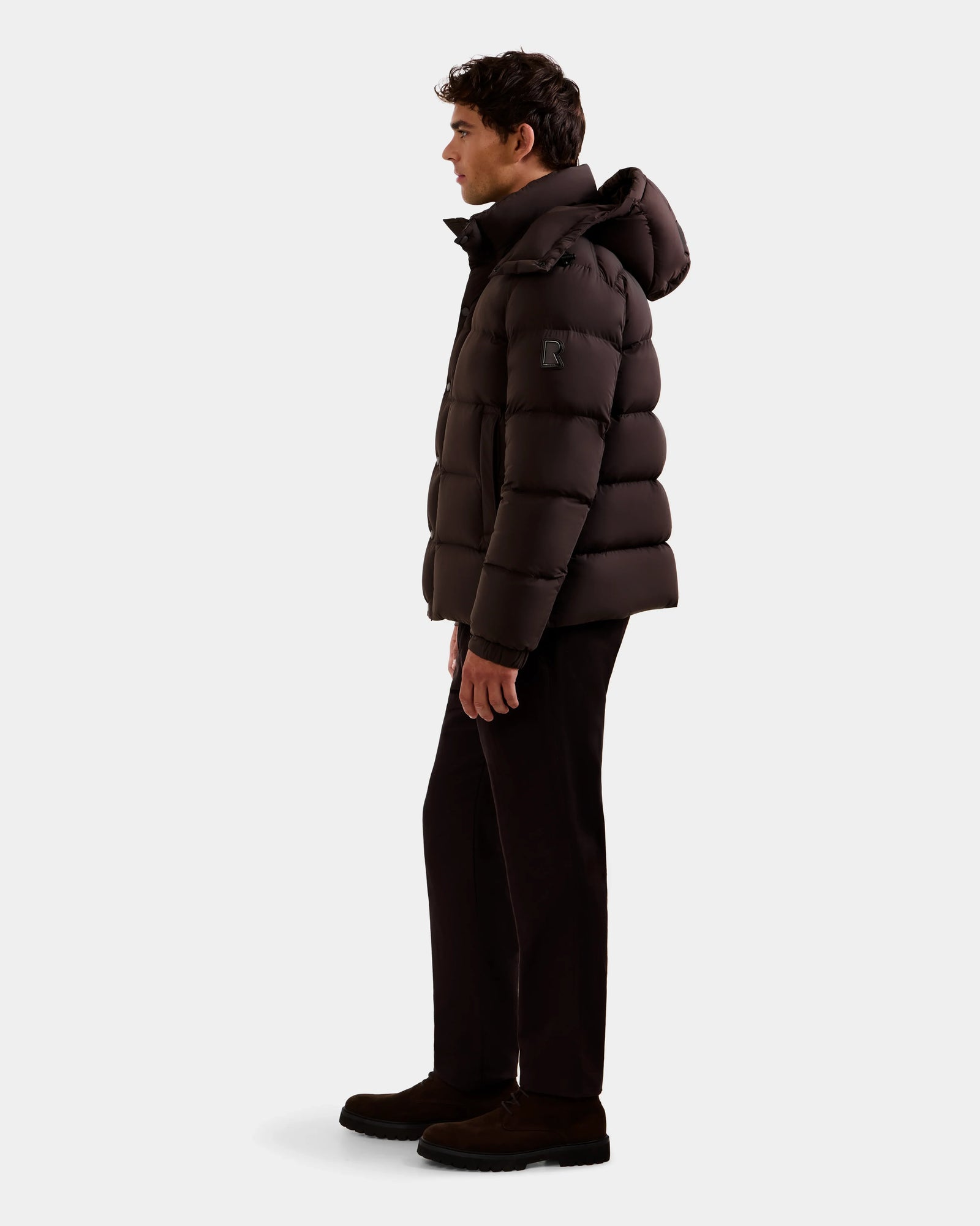 A man stands in profile wearing the Rudsak BRAYDEN M-S - M125524 Espresso men’s down puffer jacket with a hood, paired with dark pants and shoes, against a plain light background.