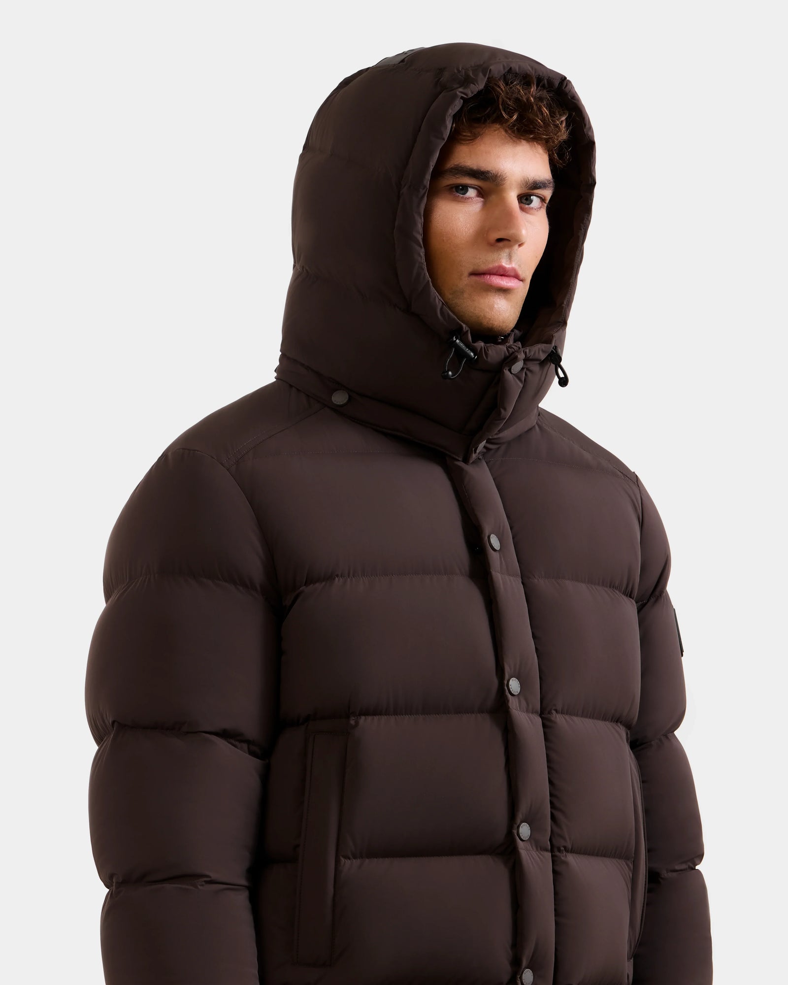 A man wearing the Rudsak BRAYDEN M-S - M125524 Espresso down puffer jacket with the hood up stands against a plain light background, looking slightly to the side with a neutral expression.