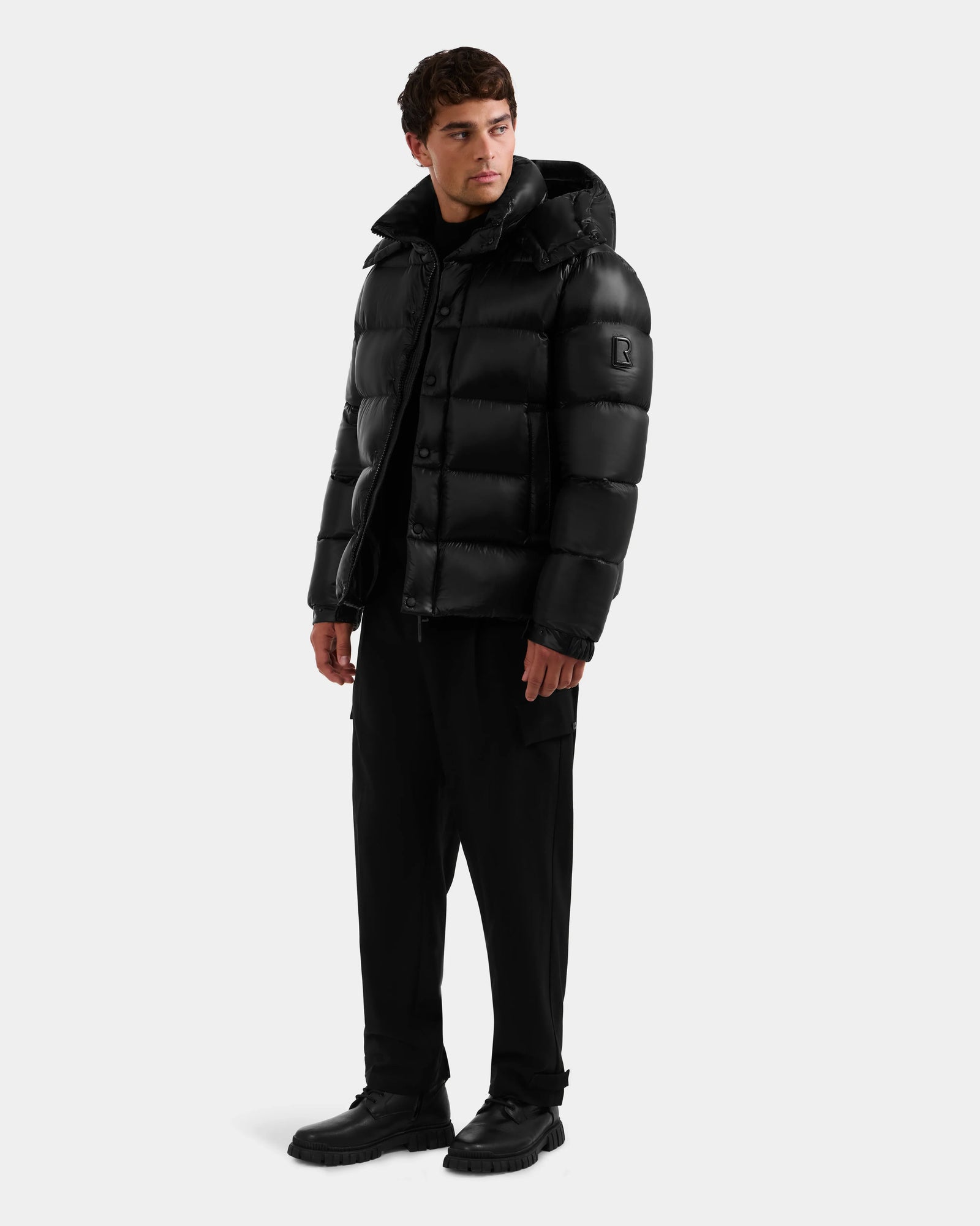 A man wears the Rudsak BRAYDEN S - M125525 Black winter jacket with a hood, paired with black pants and shoes. He stands against a light background, looking slightly left with arms relaxed at his sides.