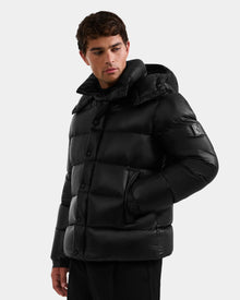 A man wears the Rudsak BRAYDEN S - M125525 Black, a shiny black quilted down puffer jacket with a hood and side pockets. He stands against a plain light background, looking down and slightly to the side in this stylish winter piece.