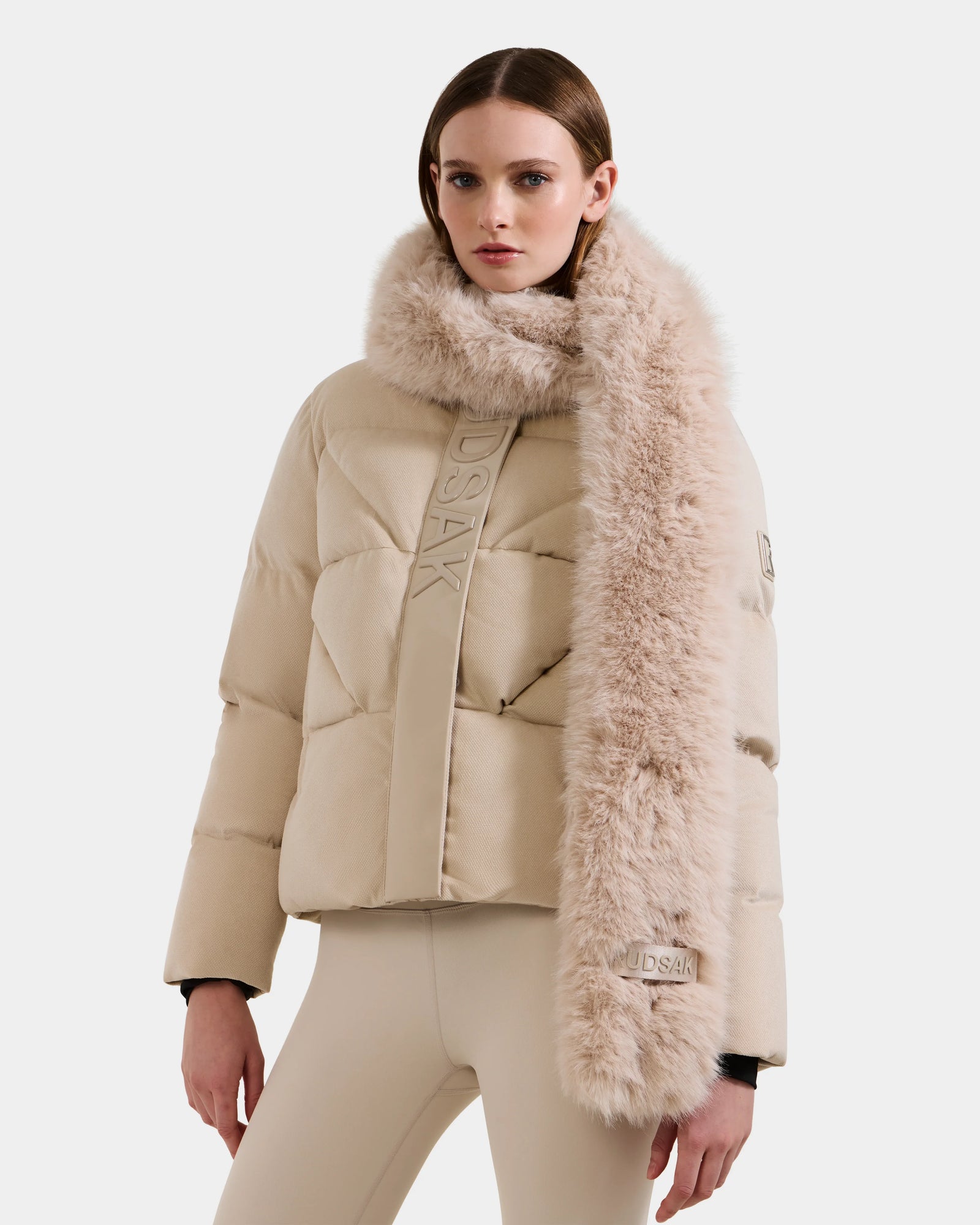 A woman wears the Rudsak CARMY - L425534 Vanilla puffer jacket with embossed lettering and a plush faux-fur scarf, paired with fitted beige pants, standing against a plain white background.