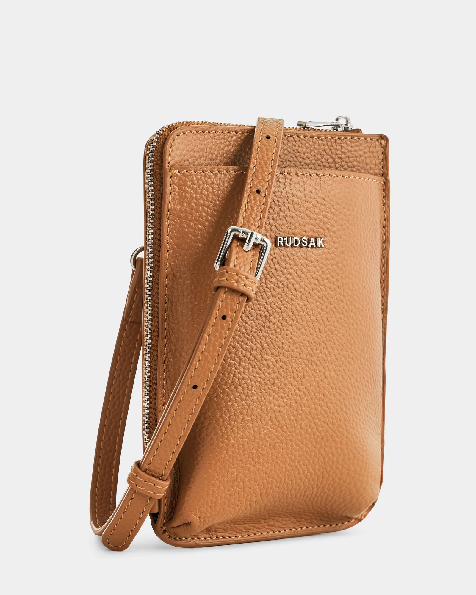 The CELYN - L325514 Taupe by Rudsak is a small, tan pebbled leather women’s crossbody bag with an adjustable strap, silver buckle, front silver RUDSAK logo, zippered top, and a minimalist modern city bag design.