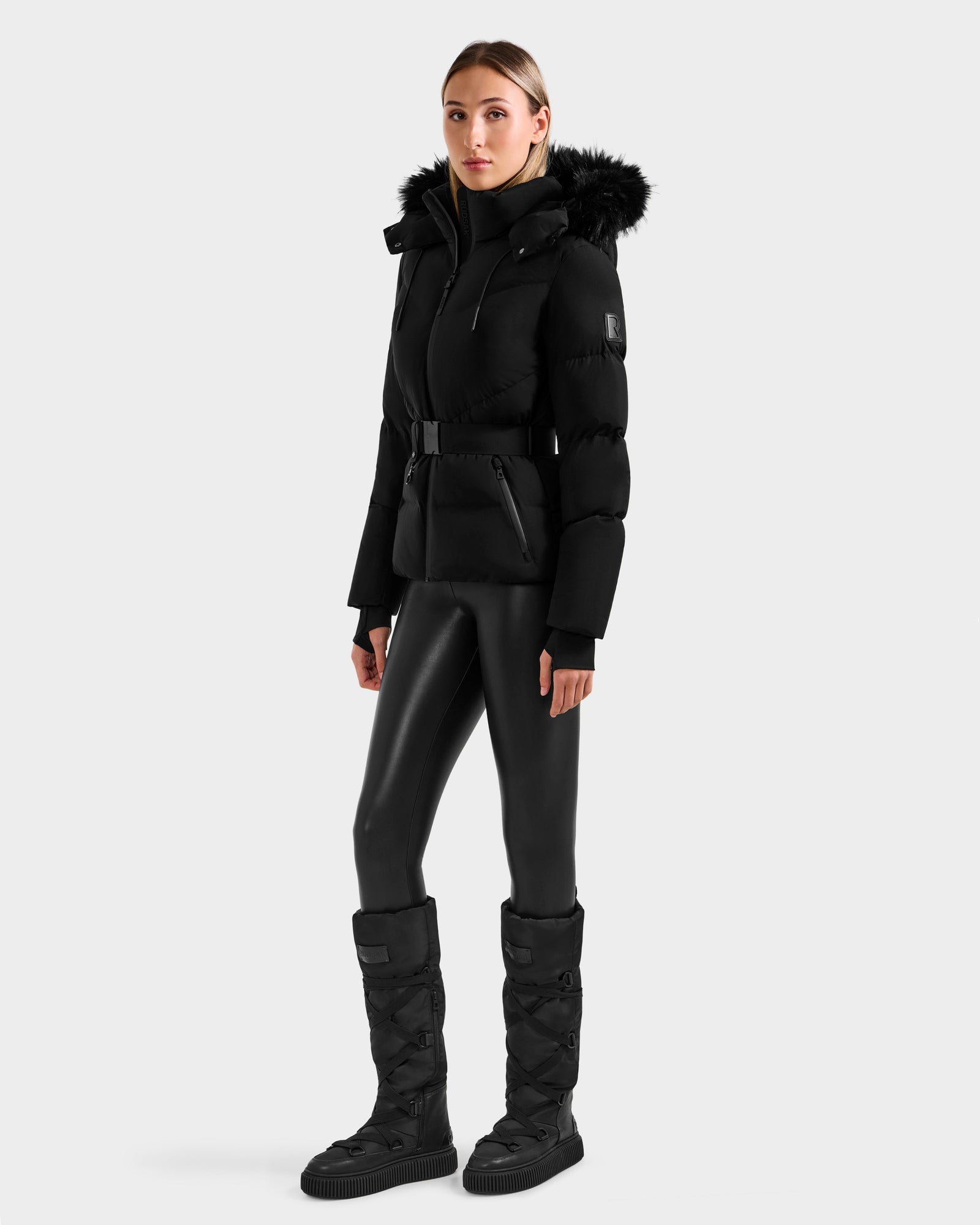 A woman stands against a light gray background wearing the Rudsak CHRISTELLE M - L125542 Black weather-resistant jacket with a fur-trimmed hood, paired with black leggings and knee-high boots.