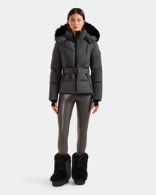 A person stands facing forward against a plain white background wearing the Rudsak CHRISTELLE M - L125542 Grey weather-resistant quilted down puffer jacket with a fur-trimmed hood.