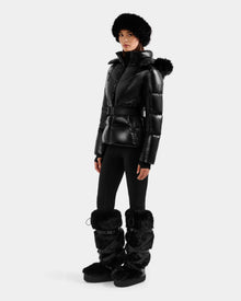 A woman wears the Rudsak CHRISTELLE S - L125544 Black down puffer jacket with a belt, paired with a black fur hat, fitted black pants, and tall faux fur-trimmed boots, standing against a plain white background.