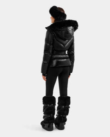 A person faces away wearing the Rudsak CHRISTELLE S - L125544 Black women’s down puffer jacket with faux fur trim, paired with fitted black pants, tall furry boots, and a fluffy headband for a chic, sporty winter look.