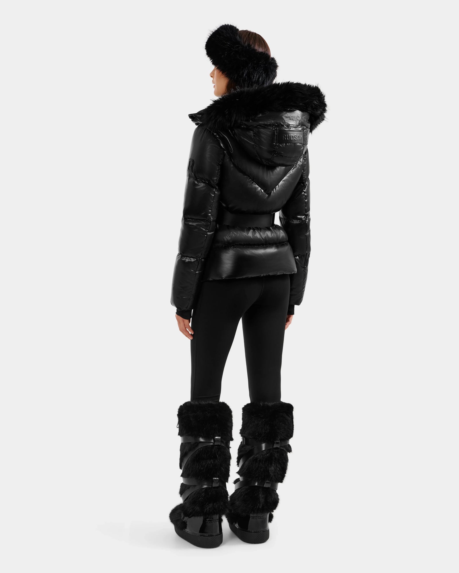 A person faces away wearing the Rudsak CHRISTELLE S - L125544 Black women’s down puffer jacket with faux fur trim, paired with fitted black pants, tall furry boots, and a fluffy headband for a chic, sporty winter look.