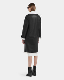 A woman with straight brown hair wears sunglasses and the Rudsak DARCY - L125539 Black vegan leather overcoat with white shearling trim, paired with black ankle boots, standing and facing away from the camera against a plain background.